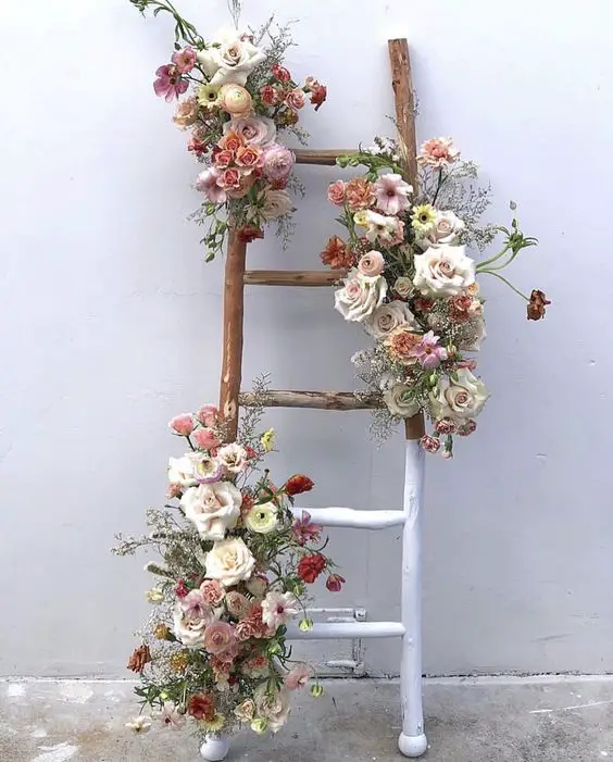 Rustic wooden ladder, half painted white, adorned with pastel flowers cascading down the sides against a white wall.