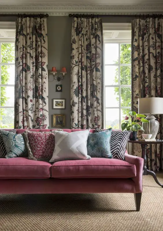 Eclectic living room with floral patterned curtains, pink velvet sofa, colorful throw pillows, and a natural fiber rug, set against gray walls with ornate molding.