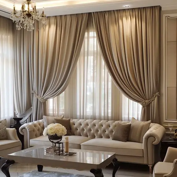 Elegant living room with layered taupe and cream curtains, a tufted cream Chesterfield sofa, crystal chandelier, and ornate coffee table, all in a neutral color palette.