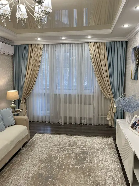 Elegant living room with layered blue and beige curtains, cream sofa, crystal chandelier, and tray ceiling, creating a serene and luxurious atmosphere.