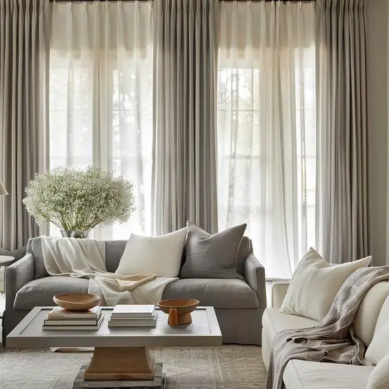 Serene living room with layered gray and white curtains, gray sofa, white armchair, modern coffee table, and a vase of white flowers, all in a neutral color palette.
