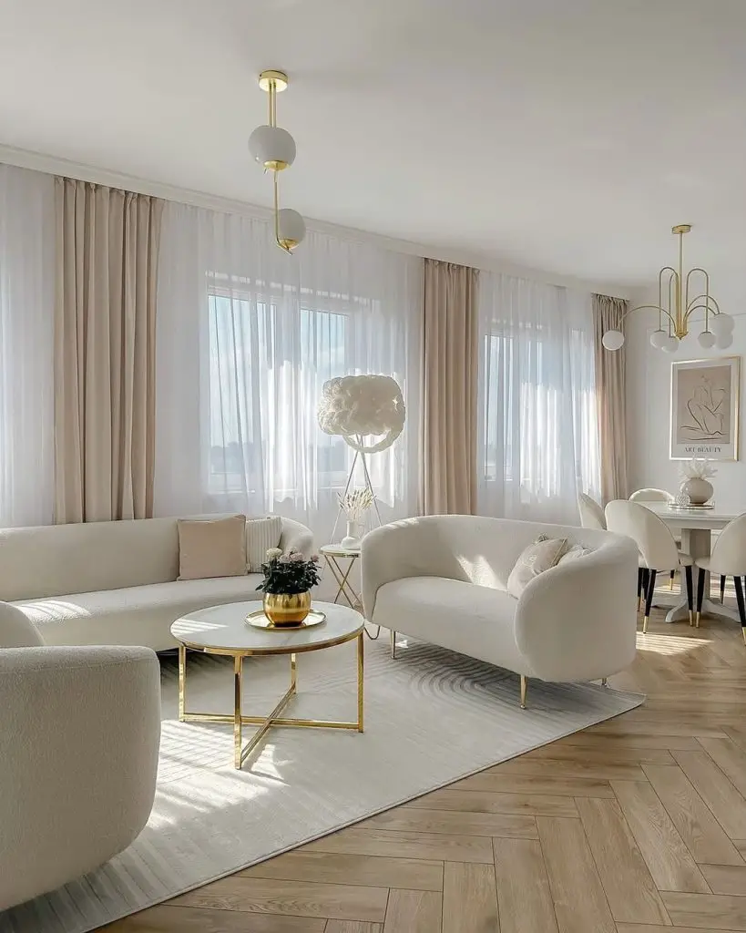 Bright living room with white furniture, gold accents, floor-to-ceiling windows, and herringbone wood flooring.