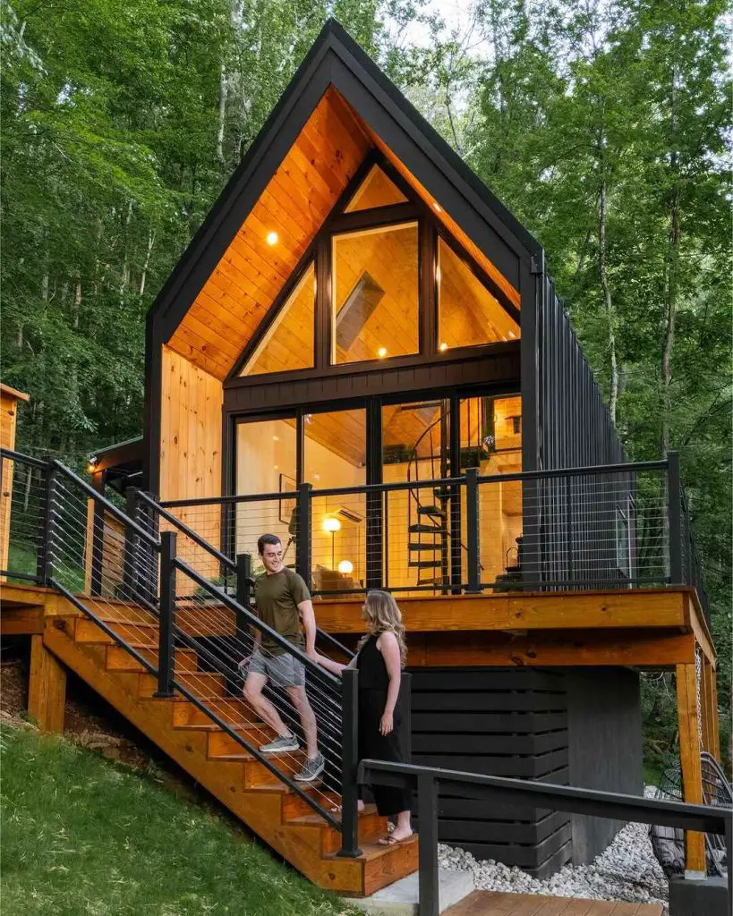 Modern A-frame cabin with large windows and a spiral staircase, surrounded by trees and greenery