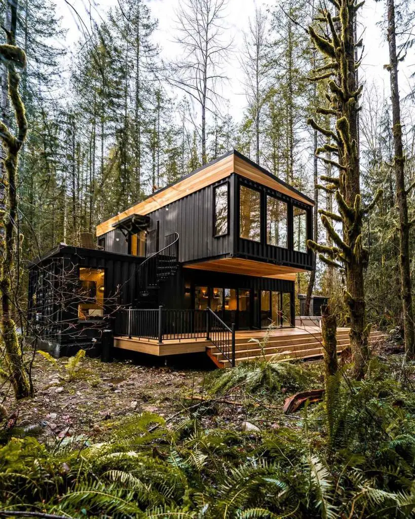 Modern black cabin with large windows and a deck, surrounded by trees and ferns