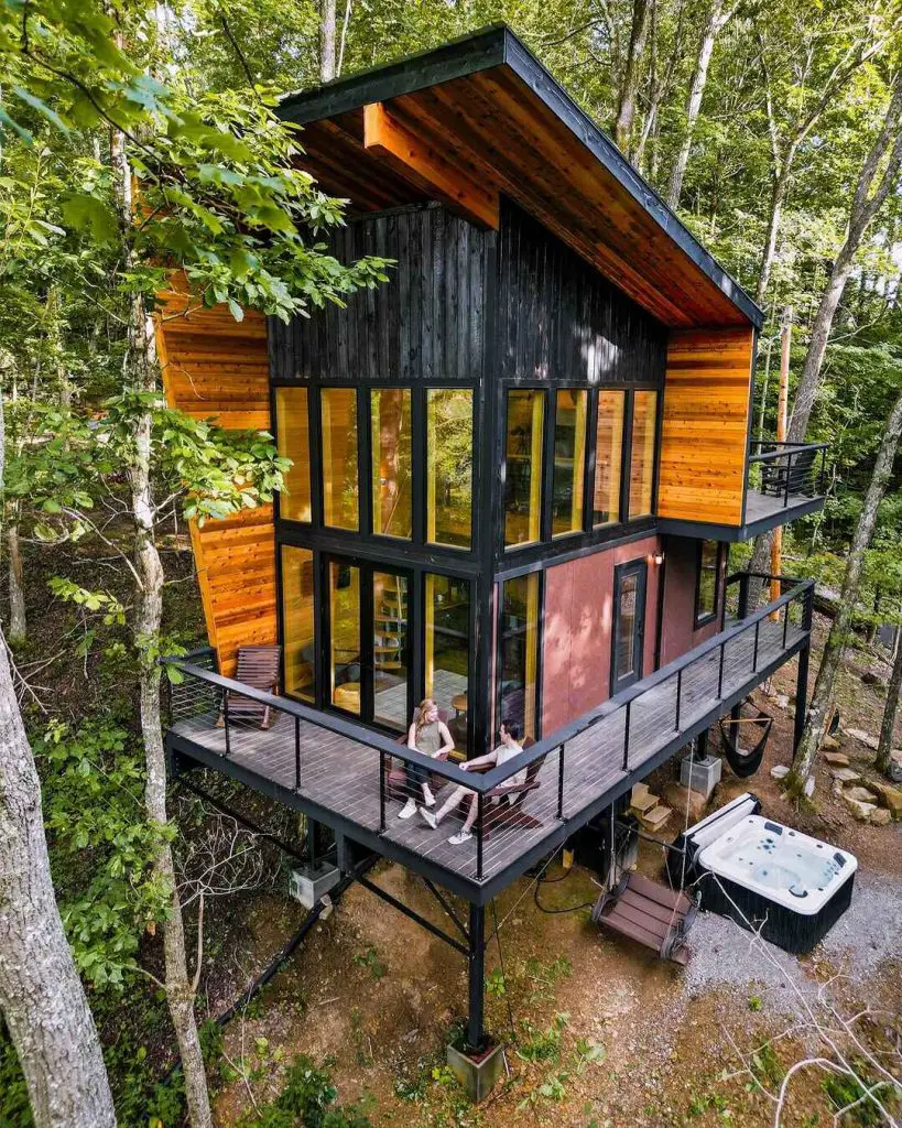 Modern treehouse cabin with large windows and a deck, surrounded by trees, featuring two people sitting outside.