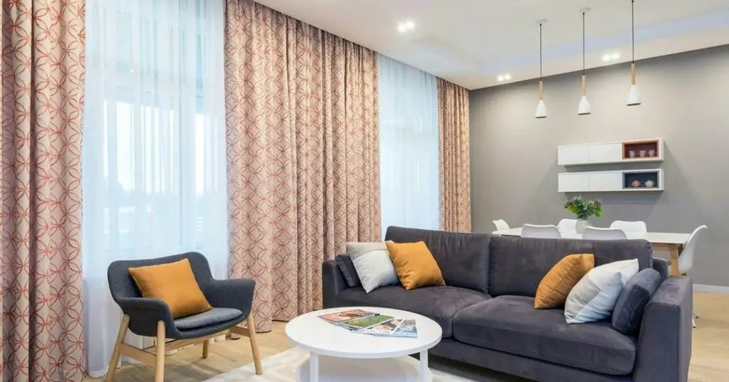 Modern living room with patterned peach curtains, gray sofa with yellow pillows, white coffee table, and pendant lights.