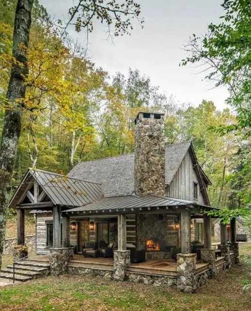 Rustic cabin made of stone and wood, featuring a large porch and chimney, surrounded by colorful autumn trees.