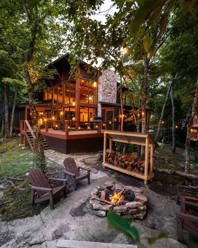Charming cabin with large windows surrounded by trees, featuring a fire pit and wooden chairs