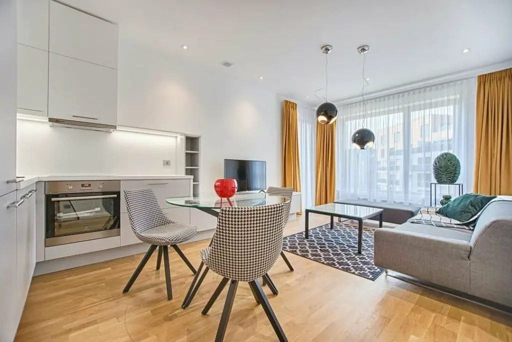 Open-plan living space with white kitchen, glass dining table with houndstooth chairs, gray sofa, and yellow curtains.