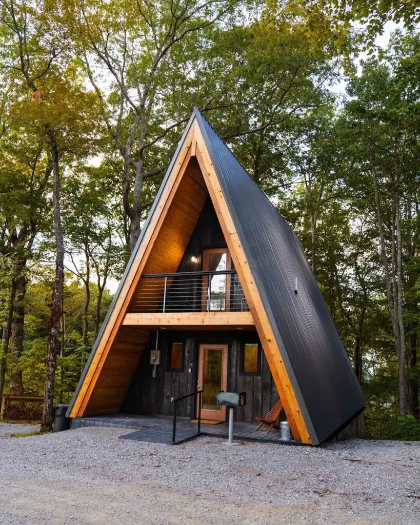 Modern A-frame cabin with wooden accents surrounded by trees and gravel path.