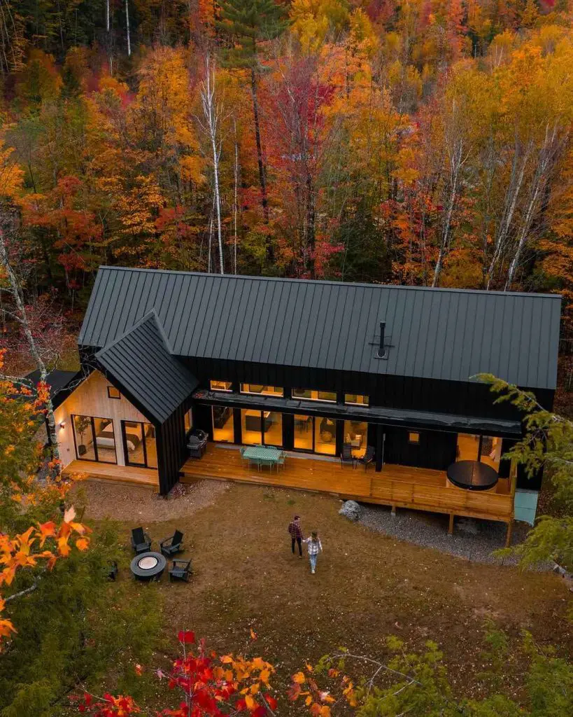 Modern cabin with large windows and a deck, surrounded by colorful autumn trees, featuring two people walking toward the entrance.