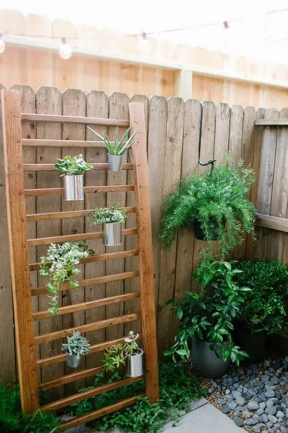 Wooden vertical plant rack with metal pots filled with greenery, leaning against a wooden fence, alongside a hanging plant and other potted plants