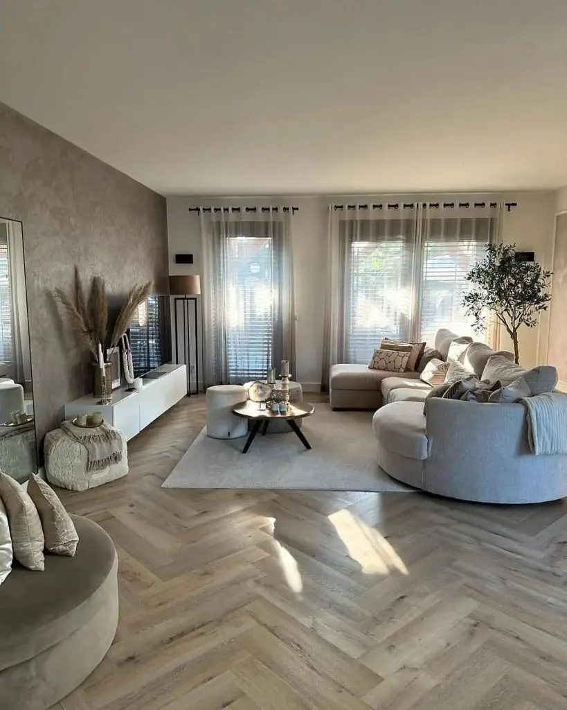 Living room with curved gray sectional, textured accent wall, herringbone flooring, and sheer curtains allowing natural light.
