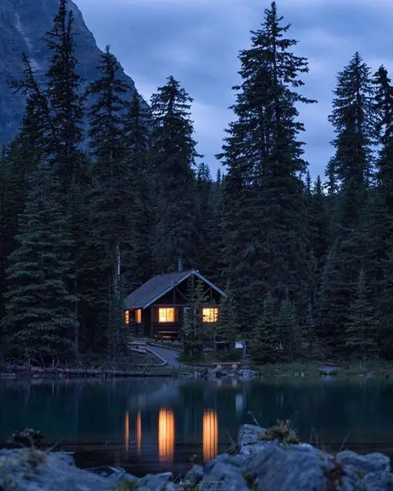Lakeside cabin surrounded by tall trees at dusk, with warm lights glowing from the windows and reflections on the water