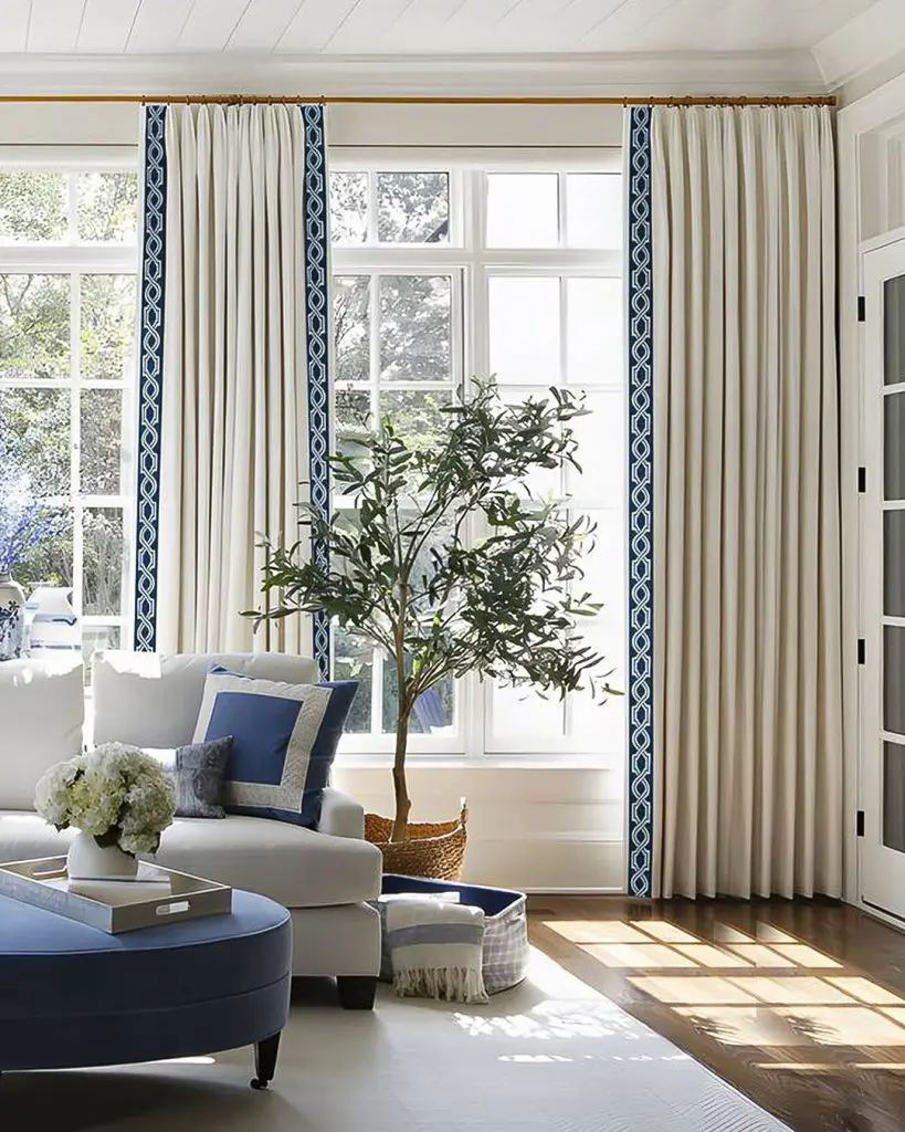 Bright living room with white curtains featuring navy blue trim, white armchair, blue ottoman, and an olive tree by large windows.