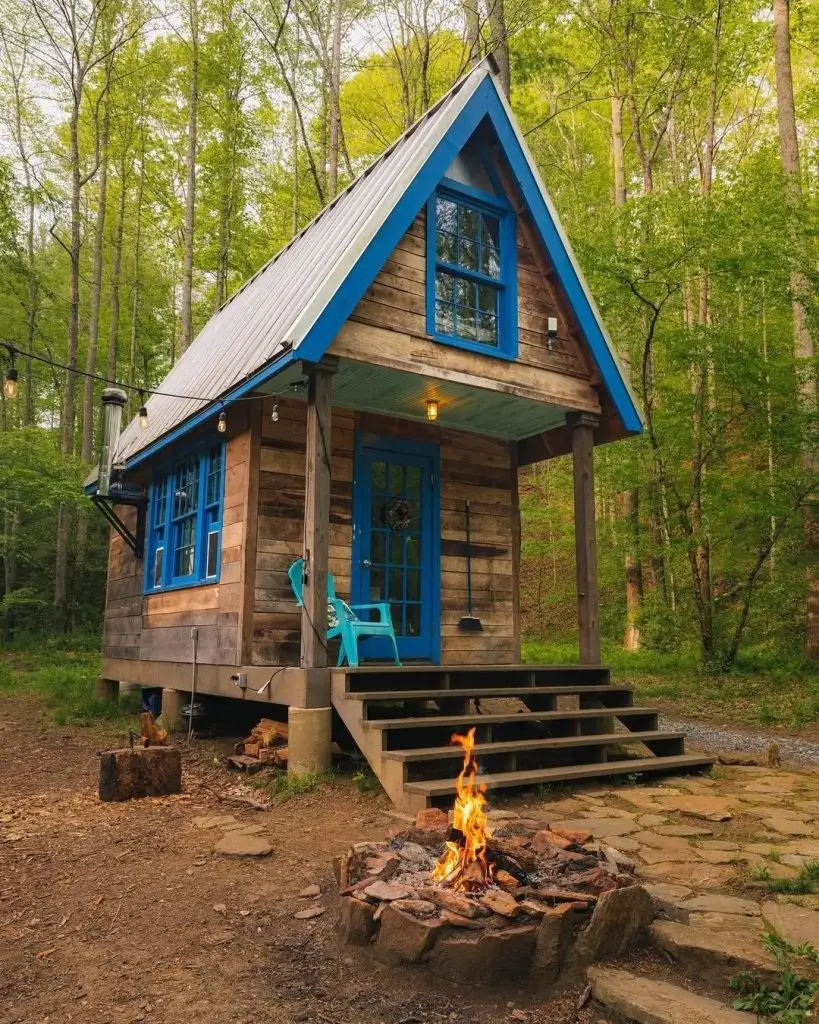 Rustic wooden A-frame cabin with bright blue trim and crackling firepit in peaceful forest setting