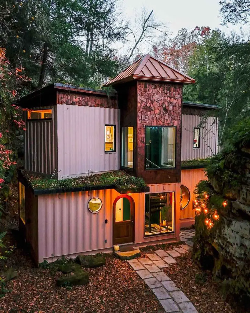 Unique cabin made from shipping containers with a copper roof, surrounded by autumn leaves and trees, featuring a stone pathway to the entrance.