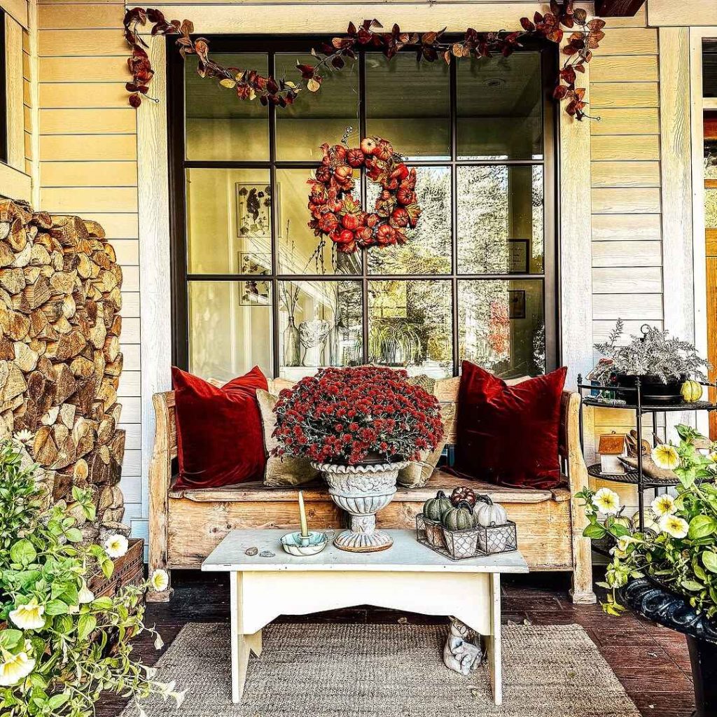 Front porch with a wooden bench, red pillows, potted mums, and autumn decor