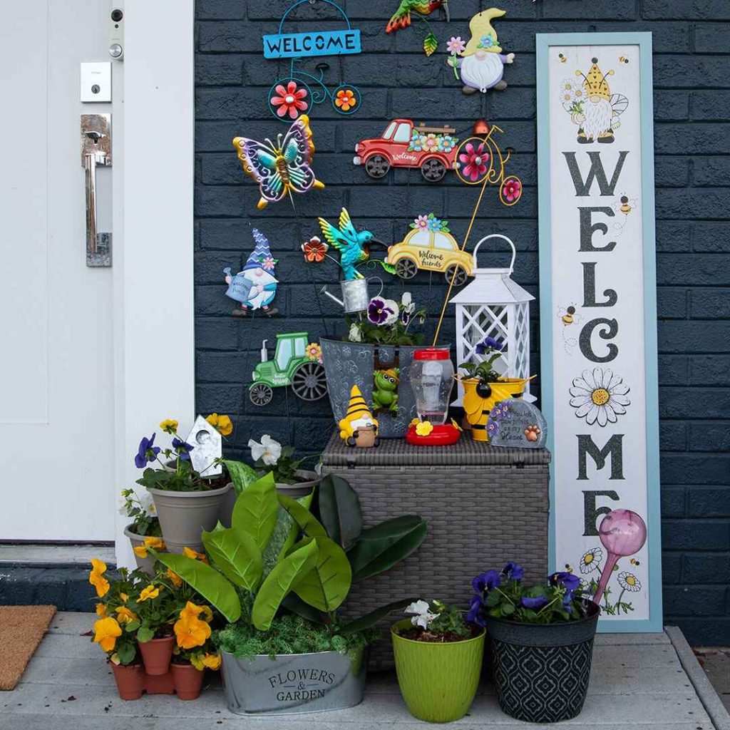 Colorful front porch garden display with potted plants, whimsical decorations, and a large welcome sign on a brick wall