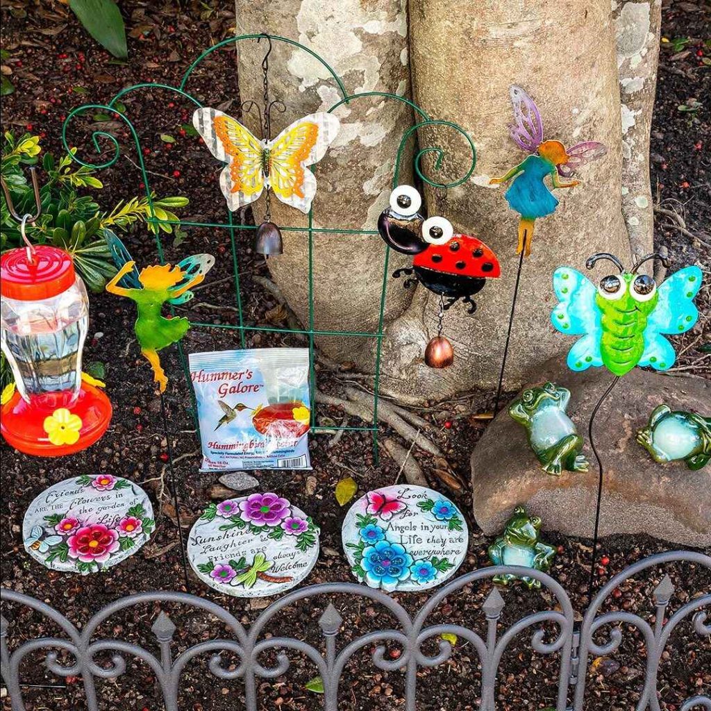Whimsical garden decor featuring colorful metal art, a hummingbird feeder, decorative stones with messages, and ceramic frogs around a tree.