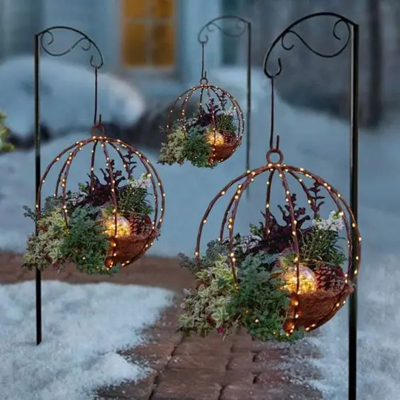 Three hanging globe-shaped planters filled with greenery and pinecones, illuminated with fairy lights, set in a snowy landscape